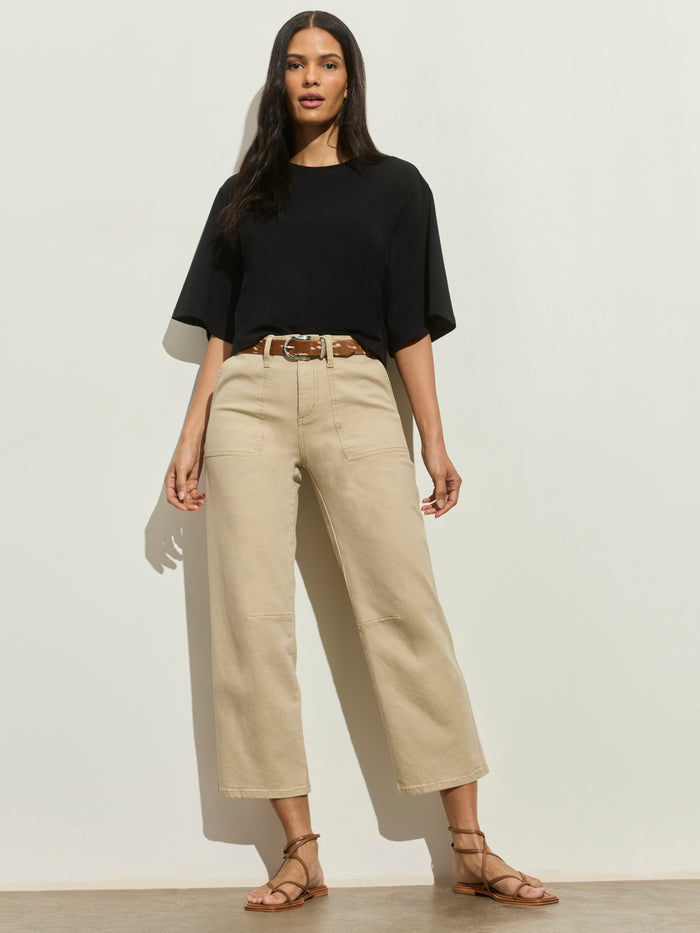 A woman with long dark hair poses against a light wall wearing Sanctuary Clothing’s foundry pant in khaki, styled with a black short-sleeve top, brown belt, and brown sandals.