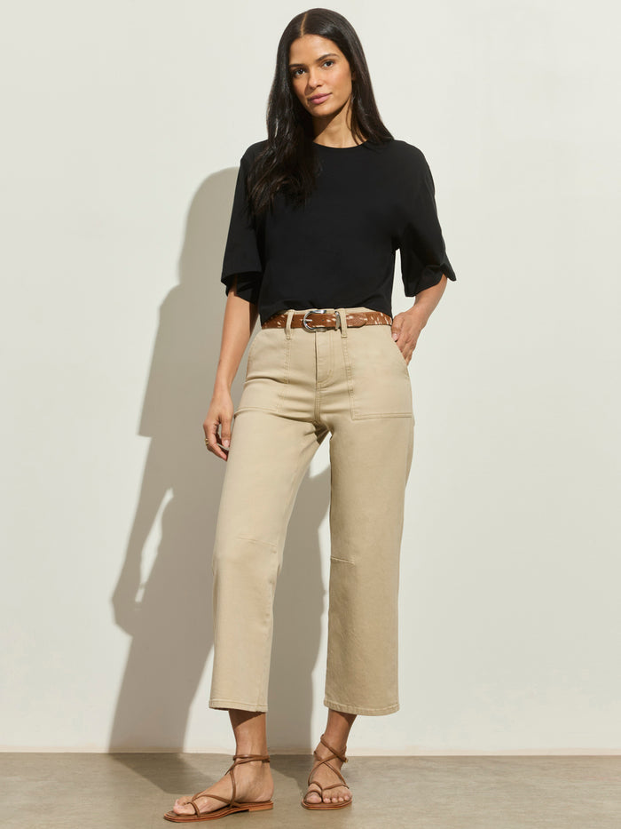 A woman with long dark hair poses confidently against a light wall, wearing Sanctuary Clothing's foundry pant khaki, a black short-sleeve top, brown belt, and tan strappy sandals.