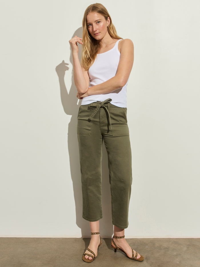 A woman stands by a light wall, wearing a white sleeveless top, Sanctuary Clothing’s foundry pant in vintage olive with a waist tie, and patterned-strap heeled sandals.