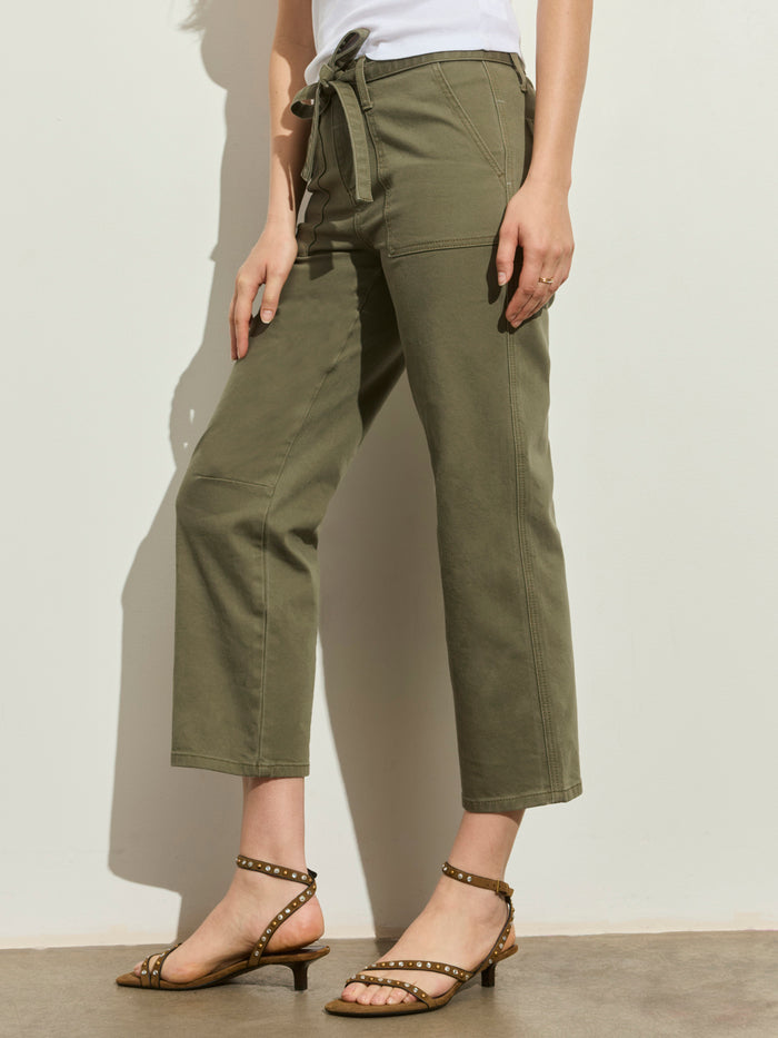 A person stands against a plain light wall wearing Sanctuary Clothing's foundry pant in vintage olive—high-waisted, wide-leg with a tie belt—paired with a white top and tan heeled sandals featuring stud details.