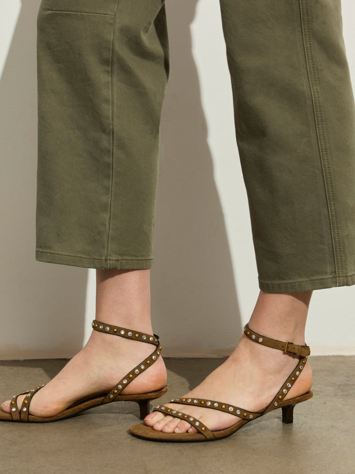 A person in Sanctuary Clothing's foundry pant in vintage olive and olive strappy sandals with small studs and low heels stands on a smooth stone floor against a light-colored wall.
