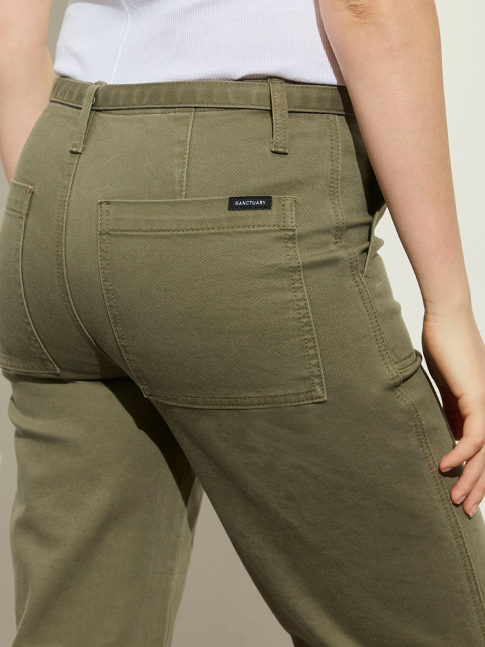 A close-up of a person wearing foundry pant vintage olive by Sanctuary Clothing, featuring a black label on the back pocket. They are also wearing a white top with their hand resting by their side.