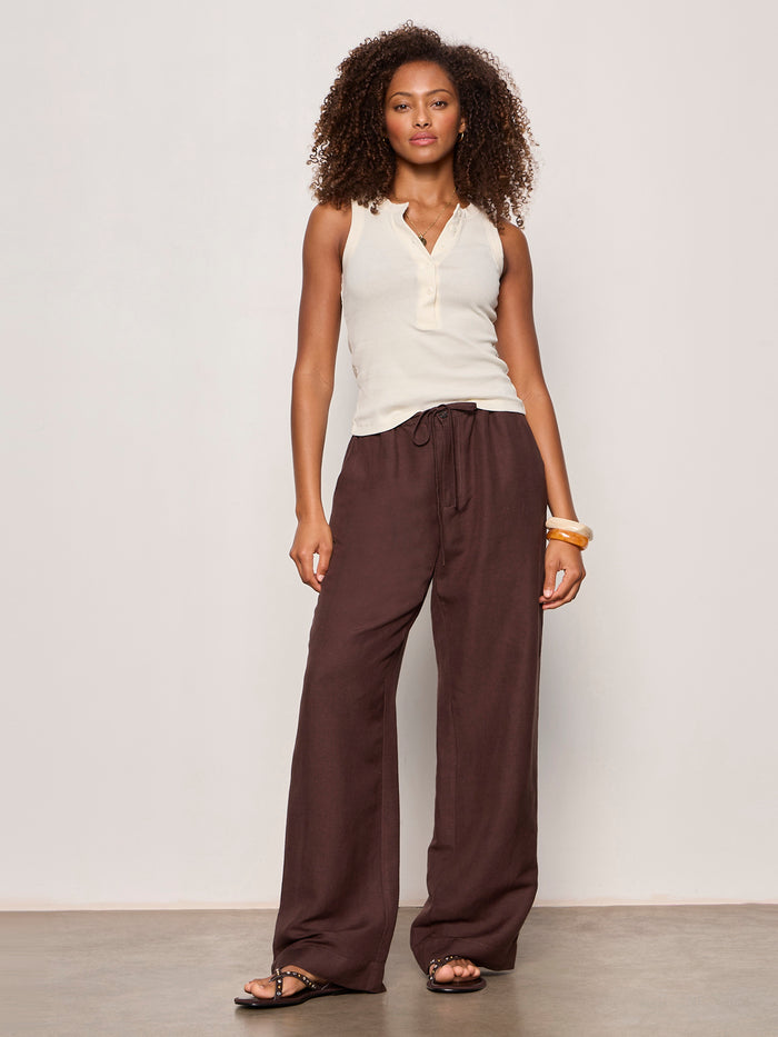 A woman with curly hair poses confidently against a plain background, wearing Sanctuary Clothing's Friday Rib Tank Collagen, loose brown drawstring pants, black sandals, and yellow bangles.