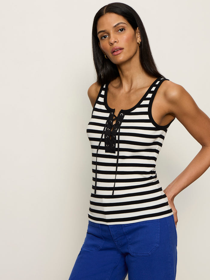 A woman with long dark hair wears the Sanctuary Clothing “go to lace up tank chalk black stripe 11,” a sleeveless, lace-up, black-and-white striped top, paired with bright blue pants against a plain light background.