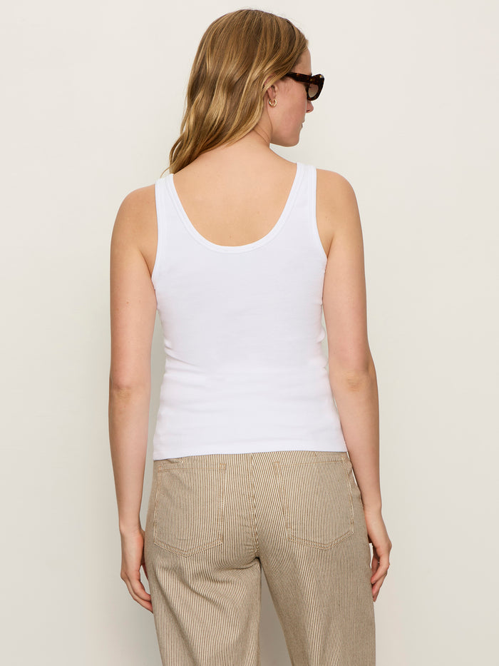 A woman with long, light brown hair wears sunglasses and the Sanctuary Clothing go to lace up tank in white, paired with beige striped pants. She stands with her back to the camera against a plain light background.