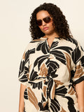 A woman with curly hair and sunglasses wears the Sanctuary Clothing good time tie shirt lotus (extended sizing), featuring a black, beige, and brown pattern, as she stands confidently against a plain light background.