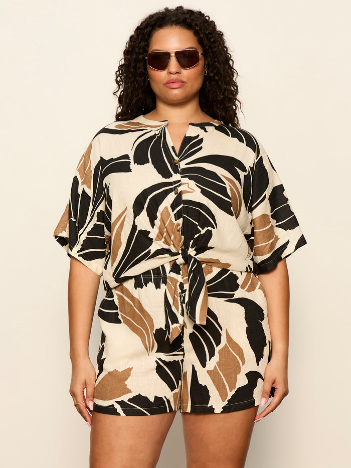 A woman with curly hair wears sunglasses and the Sanctuary Clothing good time tie shirt lotus extended sizing, a matching shorts and short-sleeve set with a bold black, brown, and beige leaf print, standing against a plain light background.
