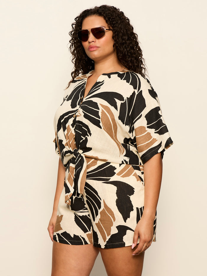 A woman with curly hair wears sunglasses and the Sanctuary Clothing good time tie shirt lotus, standing against a plain light background.