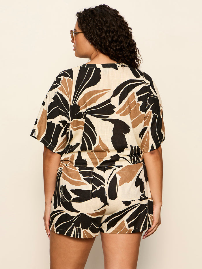 A person with curly hair and sunglasses wears the Sanctuary Clothing good time tie shirt lotus extended sizing, a short-sleeve set with bold black, tan, and cream abstract leaf print, shown from the back against a plain background.