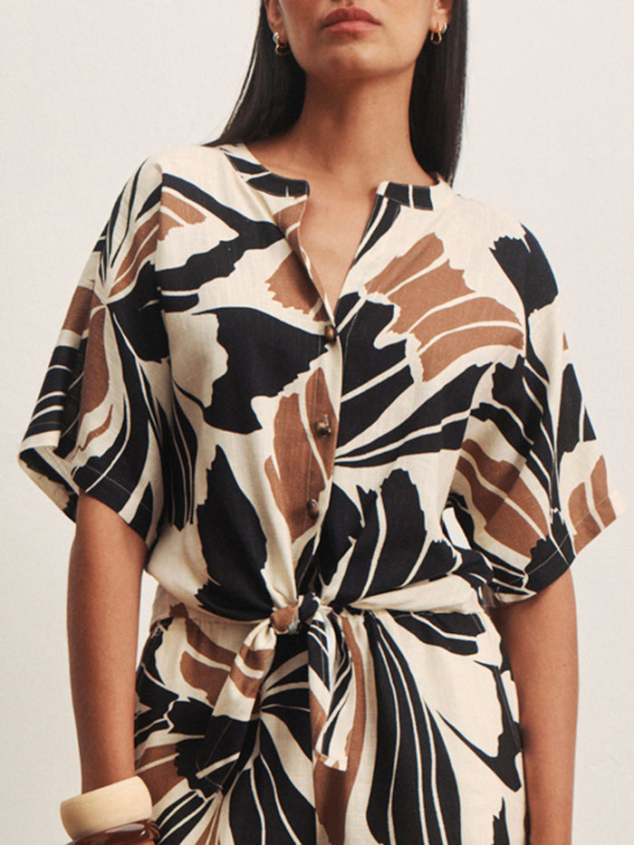 A woman wears the Sanctuary Clothing good time tie shirt lotus in petite sizing, featuring short sleeves, a button-down front, and an abstract black, brown, and white print. The dress ties at the waist; she accessorizes with a chunky bracelet.