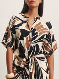 A woman wears the Sanctuary Clothing good time tie shirt lotus in petite sizing, featuring short sleeves, a button-down front, and an abstract black, brown, and white print. The dress ties at the waist; she accessorizes with a chunky bracelet.