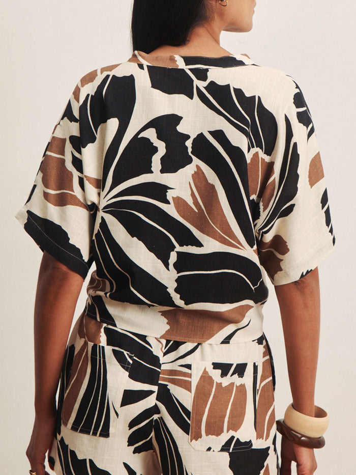 A person is shown from the back wearing Sanctuary Clothing's good time tie shirt lotus in petite sizing, featuring short sleeves and a bold abstract leaf print in black, brown, and cream. Chunky bangles also adorn their wrist.