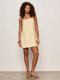 A woman with curly hair wears the Sanctuary Clothing hideaway linen tie strap dress in banana and strappy sandals, accessorized with bracelets on her right wrist, standing confidently against a plain light background.