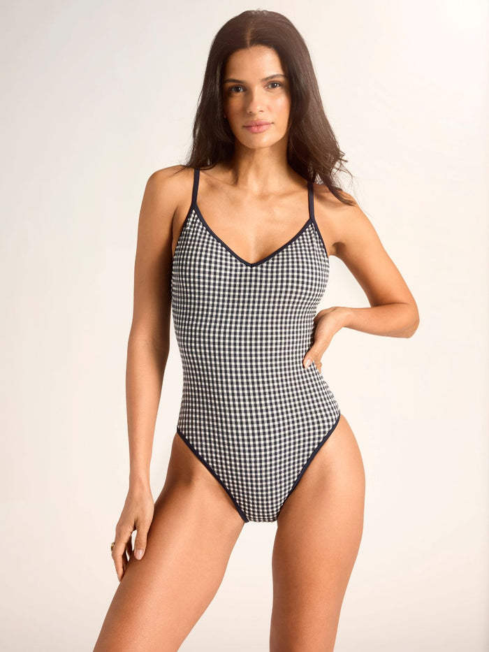 A woman poses in a Sanctuary Clothing high leg scoop neck one piece navy swimsuit with spaghetti straps, her left hand on her hip, against a plain light background.