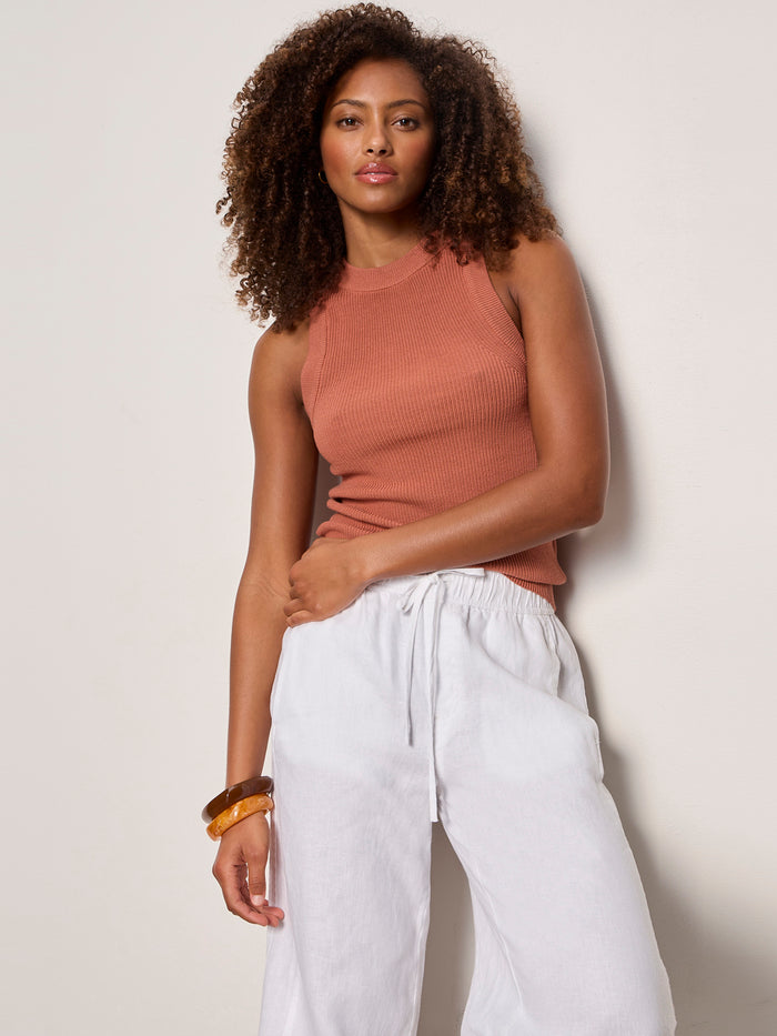A woman with curly hair wears the Sanctuary Clothing "in the mix sweater tank" in ancient clay, paired with white drawstring pants and brown bracelets, posing against a plain light background.