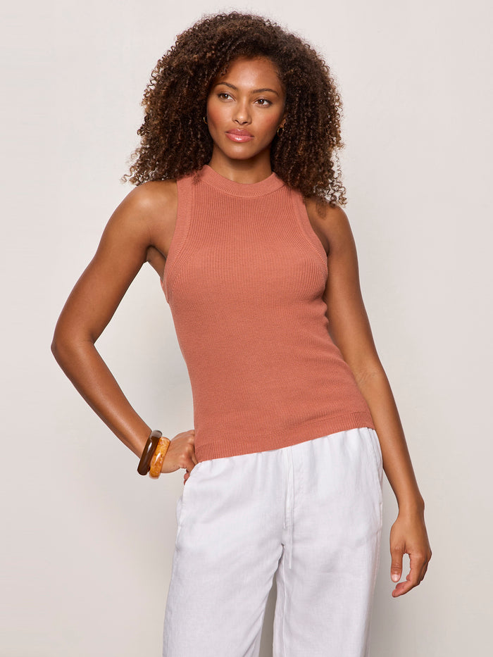 A woman with curly hair models Sanctuary Clothing's "in the mix sweater tank" in ancient clay, a sleeveless ribbed top, paired with white pants. She stands against a light background, one hand on her hip, the other relaxed by her side.