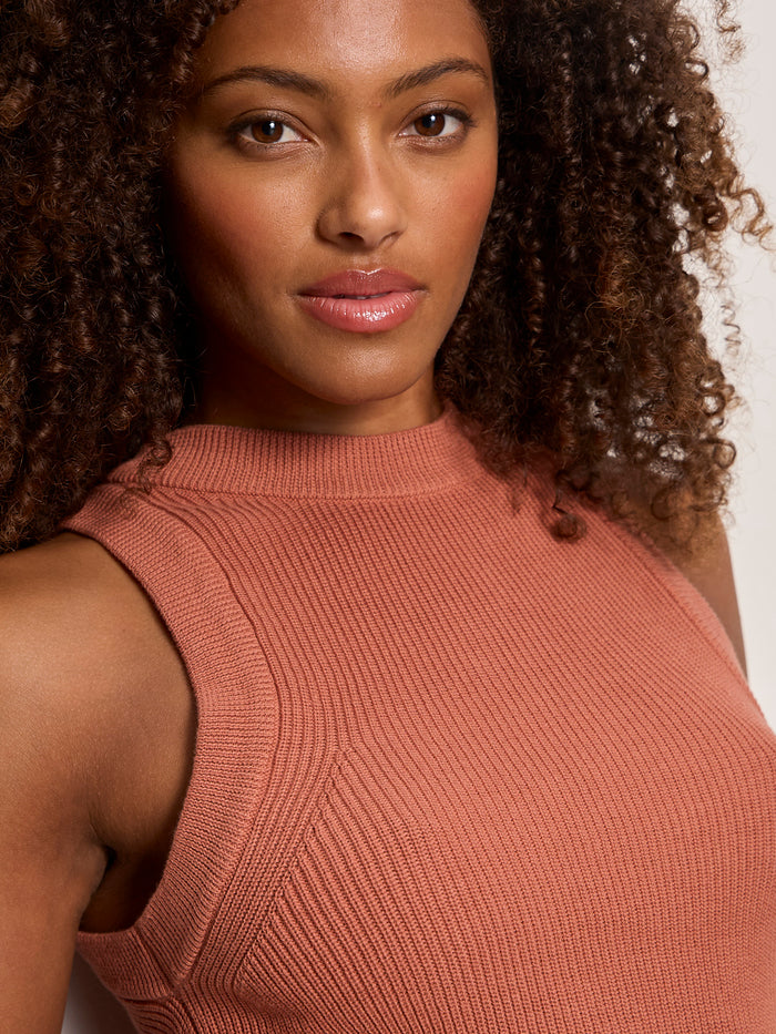 A woman with curly brown hair and glowing skin wears the Sanctuary Clothing "in the mix sweater tank" in ancient clay, confidently facing the camera against a plain, light background.