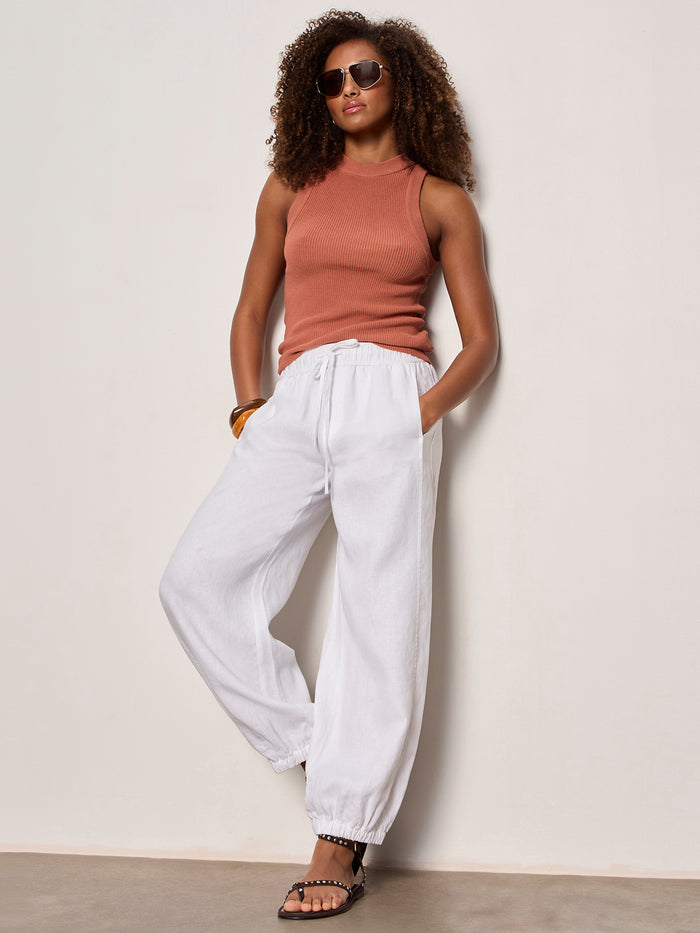 A woman wears Sanctuary Clothing's in the mix sweater tank in ancient clay, paired with white jogger pants and black sandals, standing against an off-white wall with her hands in her pockets.