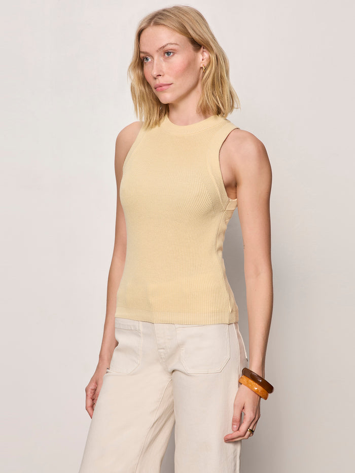 A woman with shoulder-length blonde hair wears the Sanctuary Clothing "in the mix sweater tank banana," a sleeveless, light beige ribbed top, and off-white pants. She stands against a plain background, looking slightly to the side, with two brown bangles on her right wrist.