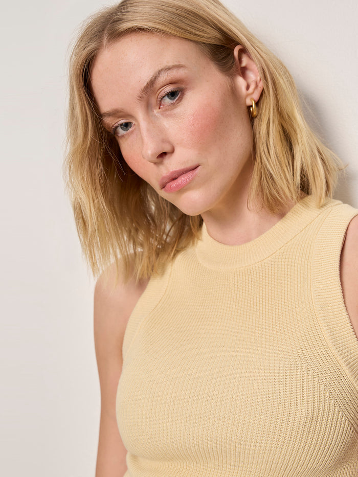 A woman with shoulder-length blonde hair wears the Sanctuary Clothing "in the mix sweater tank banana" with gold hoop earrings, standing against a light-colored wall and looking into the camera with a neutral expression.