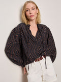 A woman with shoulder-length blonde hair wears the Sanctuary Clothing joyride relaxed volume blouse rhythmic, featuring a dark pattern, v-neck, and voluminous sleeves, styled with white high-waisted shorts and a brown belt against a light-colored wall.