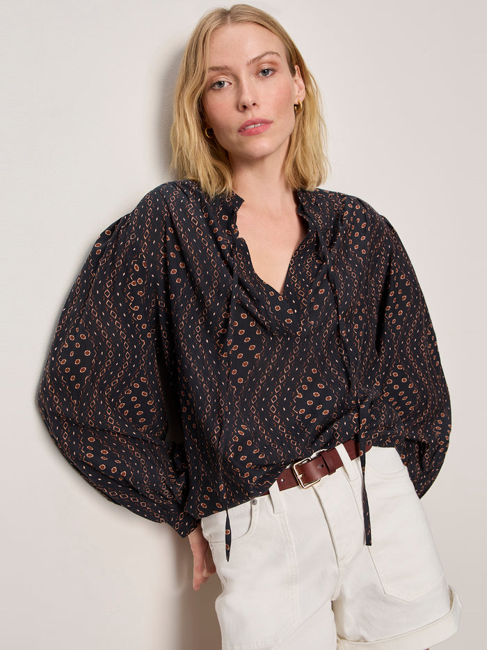 A woman with shoulder-length blonde hair wears the Sanctuary Clothing joyride relaxed volume blouse rhythmic, featuring a dark pattern, v-neck, and voluminous sleeves, styled with white high-waisted shorts and a brown belt against a light-colored wall.