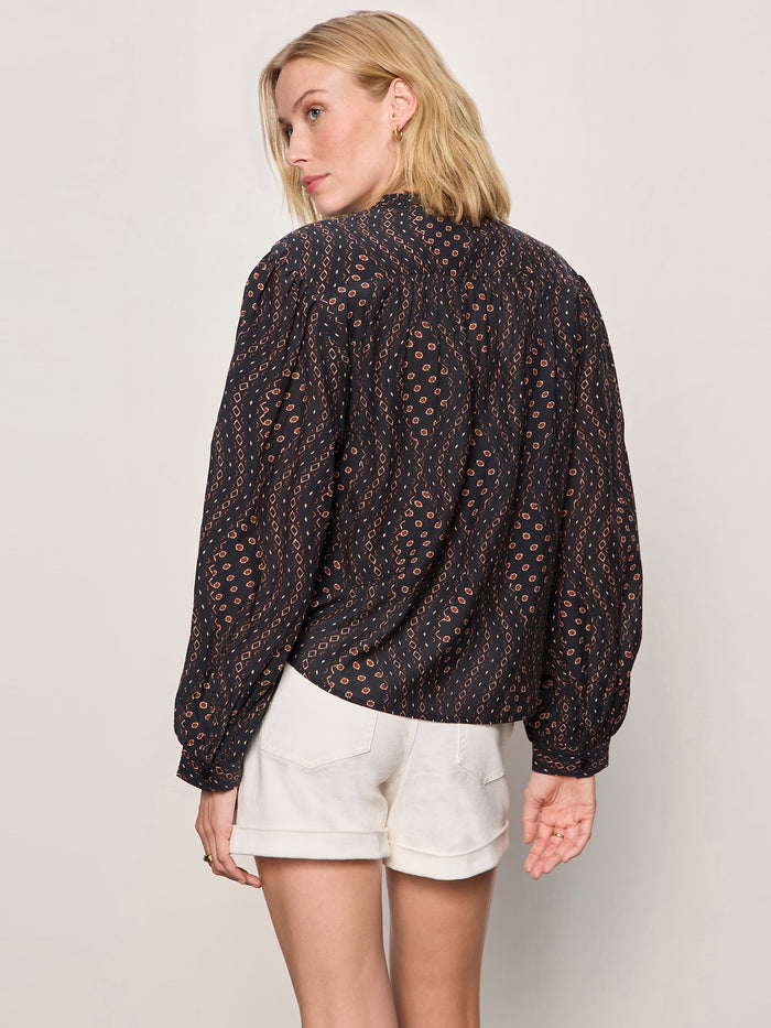 A woman with blonde hair stands facing away in Sanctuary Clothing's joyride relaxed volume blouse rhythmic, a long-sleeved black patterned top, paired with white shorts against a plain light background.