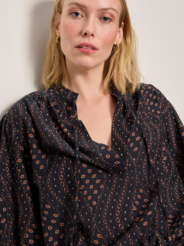 A woman with blonde hair and gold hoop earrings models Sanctuary Clothing’s joyride relaxed volume blouse rhythmic—a loose, black top with red and cream patterns—while standing against a plain, light wall and looking at the camera.