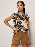 A woman wears the Sanctuary Clothing knotted muscle tee lotus, featuring a black, tan, and white floral print with rolled sleeves, paired with tan high-waisted shorts. She stands against a light background with one hand on her hip.