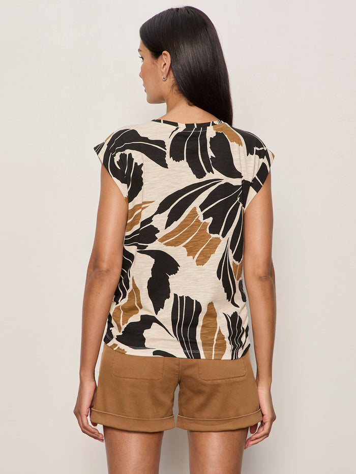 A woman with long dark hair stands facing away from the camera against a plain light background, wearing Sanctuary Clothing's knotted muscle tee lotus in beige with bold black and brown leaf patterns, paired with brown shorts.