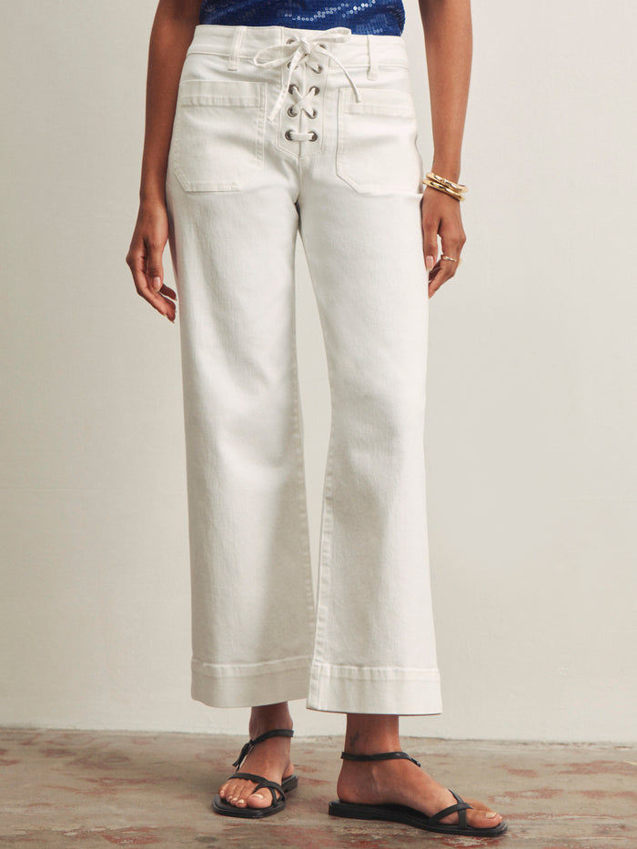 A person wearing Sanctuary Clothing's lace up mariner chalk high-waisted, wide-leg white jeans with front patch pockets, paired with black sandals, gold bangles, and a blue top stands against a neutral background.
