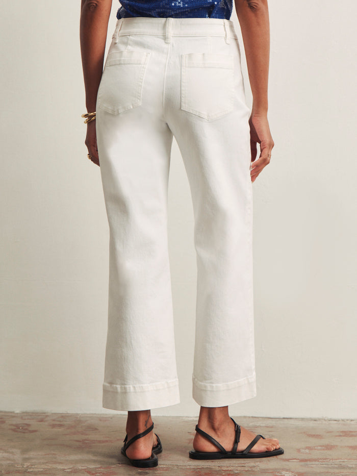 A person stands facing away, wearing Sanctuary Clothing’s lace up mariner chalk wide-leg jeans with cuffed hems, a dark blue top, black strappy sandals, and gold bracelets against a plain neutral background.