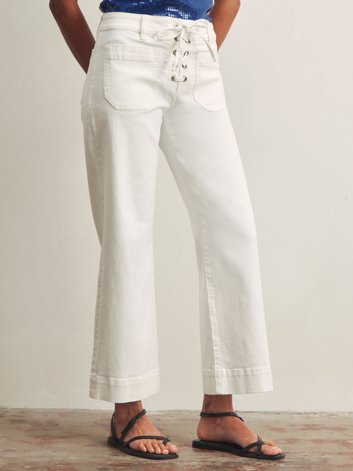 A person wears Sanctuary Clothing's lace up mariner chalk high-waisted, wide-leg white pants with a lace-up front and front pockets, paired with black strappy sandals, standing against a plain light-colored wall.
