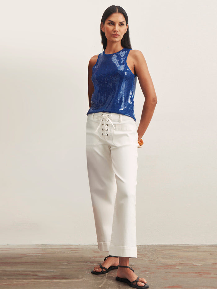 A woman with straight dark hair and a neutral expression wears the Sanctuary Clothing "lace up mariner chalk" high-waisted white wide-leg pants with front lace-up detail, paired with a sleeveless blue sequin top and black sandals, against a plain background.