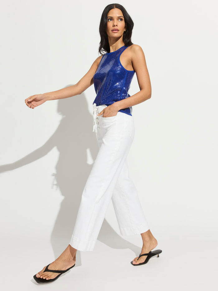 A woman walks confidently in Sanctuary Clothing's lace up mariner chalk pants, a shiny sleeveless blue top, and black flip-flop sandals against a plain white background.