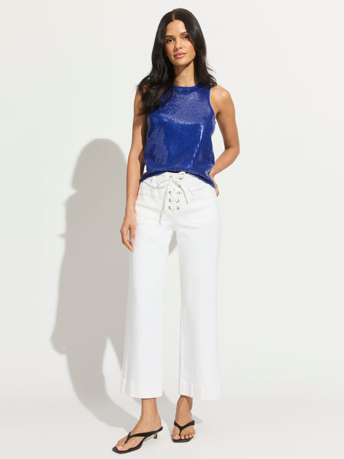 A woman with long dark hair stands against a white background wearing Sanctuary Clothing’s high-waisted lace up mariner chalk wide-leg pants, a sleeveless blue sequin top, and black heeled sandals, with her hands in her pockets.
