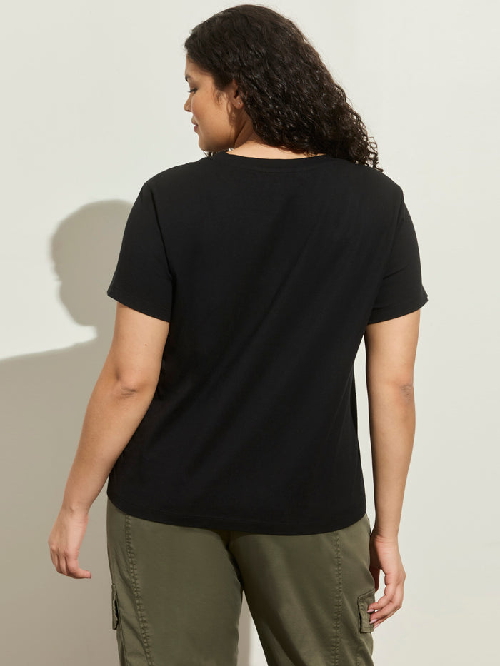 A person with curly dark hair stands with their back to the camera, wearing the Sanctuary Clothing lace up timeless tee in black (extended sizing) and olive green cargo pants against a light background.