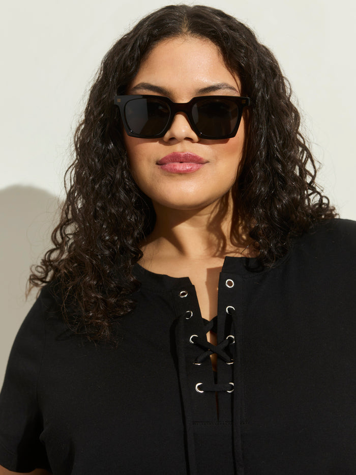 A woman with curly dark hair wears black sunglasses and the Sanctuary Clothing lace up timeless tee in black (extended sizing), standing against a plain light background.