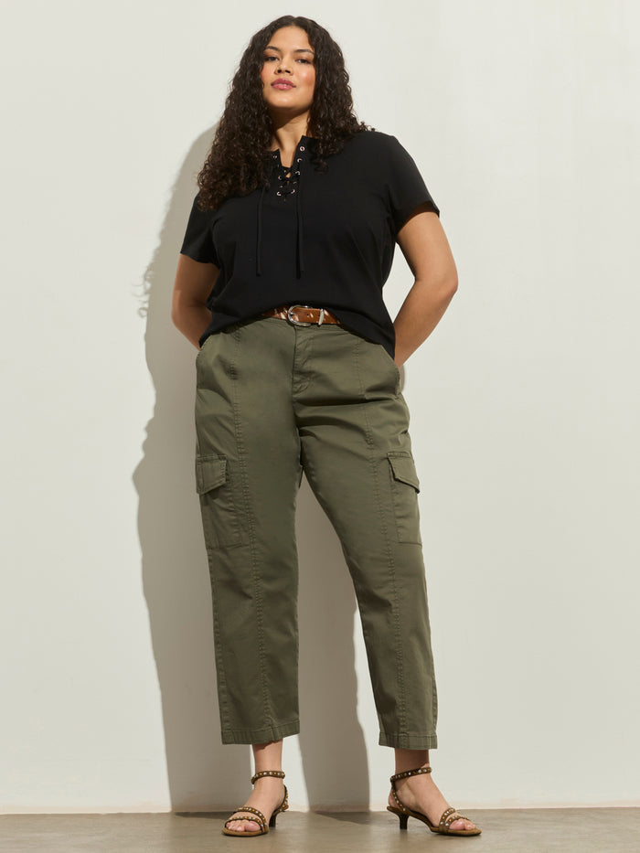 A woman with curly hair confidently poses against a light wall, wearing the Sanctuary Clothing lace up timeless tee black extended sizing, olive cargo pants with pockets, a brown belt, and sandals.