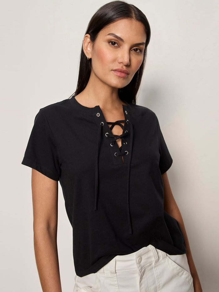 A woman with long dark hair models the Sanctuary Clothing lace up timeless tee in black, paired with light-colored pants, standing against a plain light background and gazing confidently at the camera.
