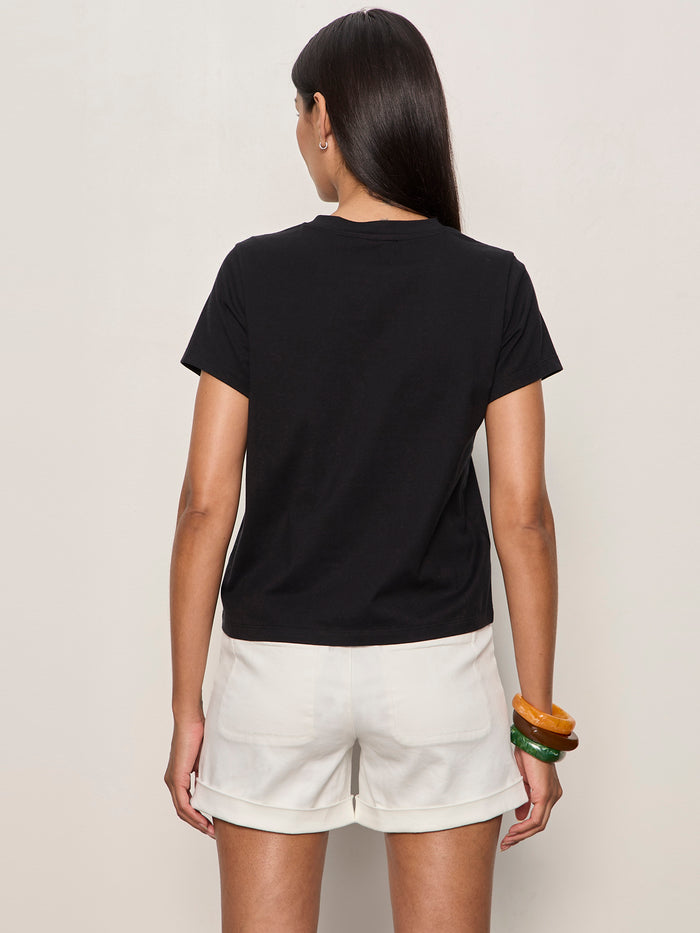 Wearing the Sanctuary Clothing lace up timeless tee in black, a person with long dark hair and white shorts stands facing away from the camera against a plain light wall, with bracelets on their right wrist.