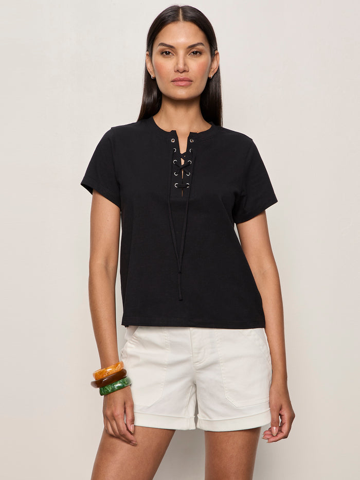 A woman with straight dark hair wears the Sanctuary Clothing lace up timeless tee in black with white shorts and colorful bangles on her left wrist, standing against a plain light background.