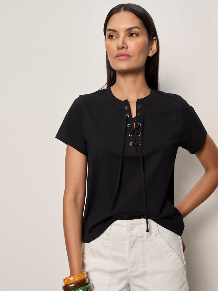 A woman with long dark hair wears the Sanctuary Clothing lace up timeless tee in black and white pants, standing against a light wall. She has one hand in her pocket and colorful bangles on her wrist.