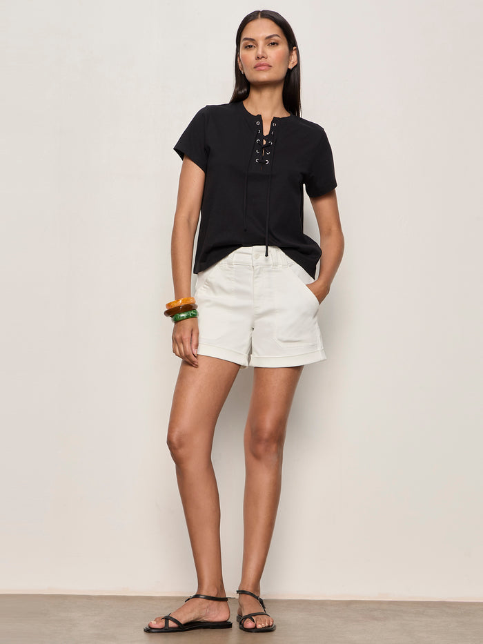A woman with long dark hair stands against a plain wall wearing the Sanctuary Clothing lace up timeless tee in black, paired with white shorts, black sandals, and orange bangles on her left wrist. She has a neutral expression.