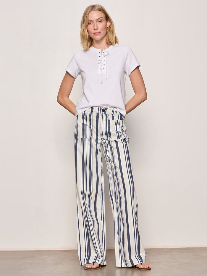 A woman stands barefoot against a plain background in the Sanctuary Clothing lace up timeless tee in white and high-waisted, wide-leg navy and white striped pants, with her hands in her pockets.