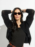 A woman with long dark hair and black sunglasses poses with arms raised, wearing the Sanctuary Clothing laguna openwork bomber in black over a black top, against a plain white background.