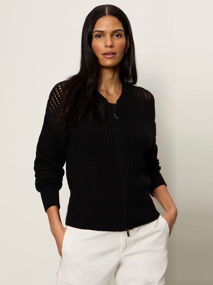 A woman with long dark hair wears the Sanctuary Clothing “laguna openwork bomber” in black and white pants, standing against a plain light background with her hands in her pockets.