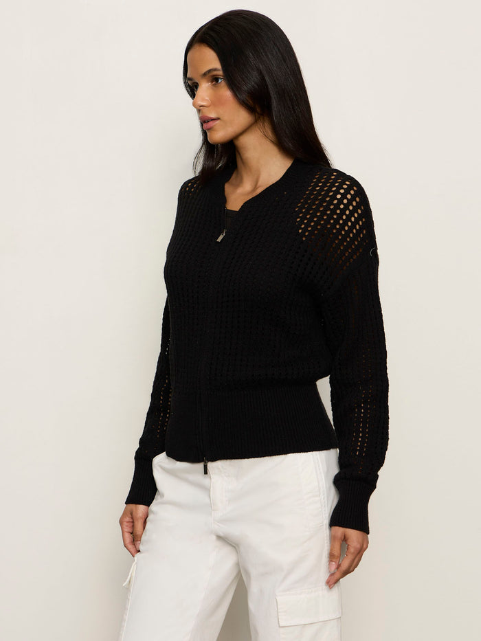A woman with long dark hair models Sanctuary Clothing's laguna openwork bomber in black, featuring mesh sleeve and shoulder details, paired with white pants against a plain light background.