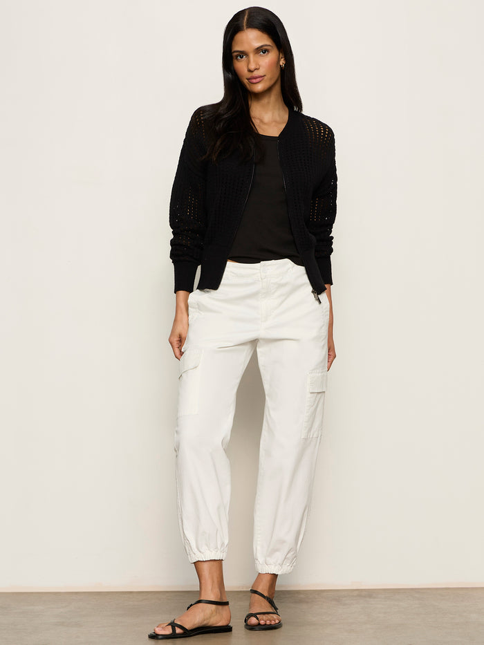 A woman with long dark hair stands against a plain background wearing the Sanctuary Clothing laguna openwork bomber in black over a black top, paired with white cargo pants and black sandals, posing relaxed with one hand in her pocket.