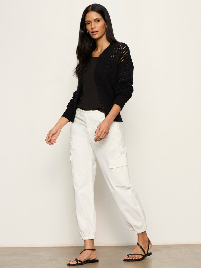 A woman with long dark hair stands against a plain background wearing the Sanctuary Clothing laguna openwork bomber in black, paired with a black top, white cargo jogger pants, and black sandals. She looks slightly to the side with a relaxed expression.